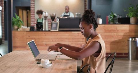 Remote worker typing at cafe table with laptop, coffee, smartwatch in industrial brick cafe