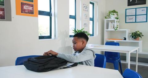 African American Boy Searching Backpack While Sitting in Bright Classroom