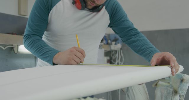 Caucasian man measures a surfboard in a workshop. He's focused on precision while crafting custom surf equipment.