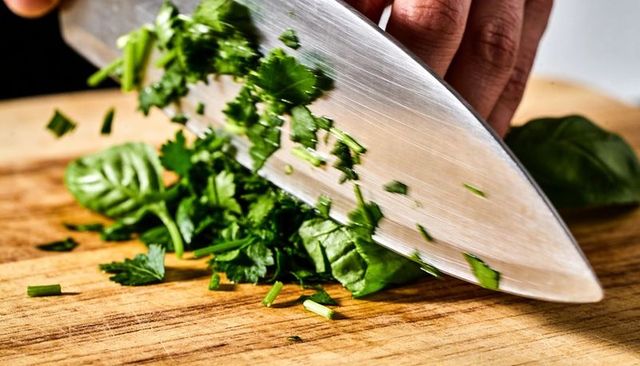 Chef knife chopping fresh herbs on wooden cutting board, hand guiding blade close-up macro