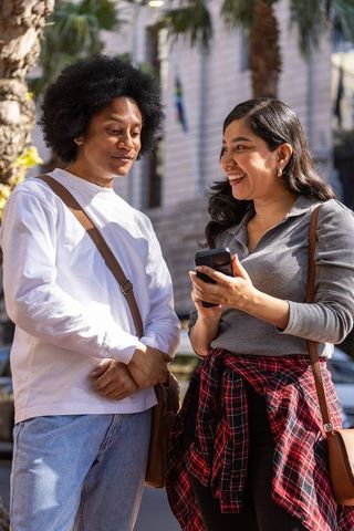 Sunlit urban duo sharing smartphone and laughter on palm-lined city sidewalk