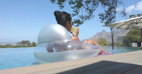 Mature woman relaxing on silver float in infinity pool sipping cocktail with mountain view
