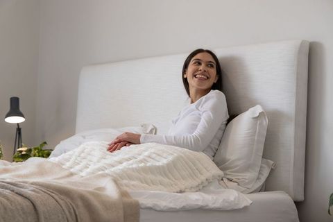 Smiling woman sitting up in cozy neutral bedroom with textured blanket and tall headboard