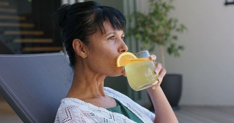 Woman sipping lemon drink while relaxing on terrace chair, sunlit summer refreshment
