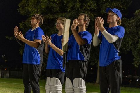 Night cricket team clapping on grass outfield in blue jerseys holding bat and pads