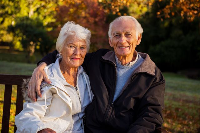Elderly couple sitting on a bench in a park during autumn, smiling and enjoying each other's company. Ideal for use in advertisements for retirement communities, healthcare services, senior lifestyle magazines, and family-oriented campaigns. Highlights themes of love, companionship, and peaceful retirement.