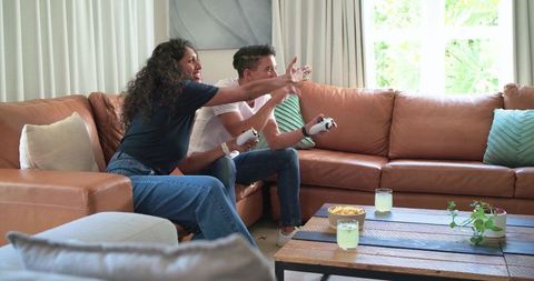 African couple playing video game on leather sofa with controllers, snacks and drinks