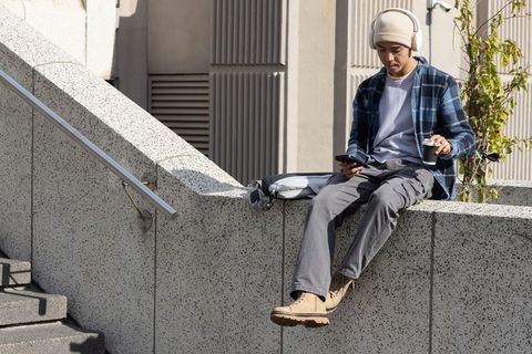 Asian commuter sitting on concrete ledge listening to music and checking phone with coffee