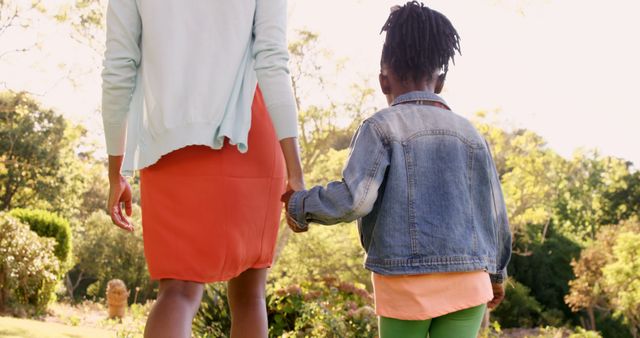 African American mother and daughter holding hands, walking in park. Mother has dark hair, wearing a red skirt; child in a denim jacket and green pants