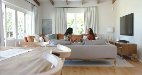 Two friends chatting on neutral sofa sharing coffee in sunlit modern living room