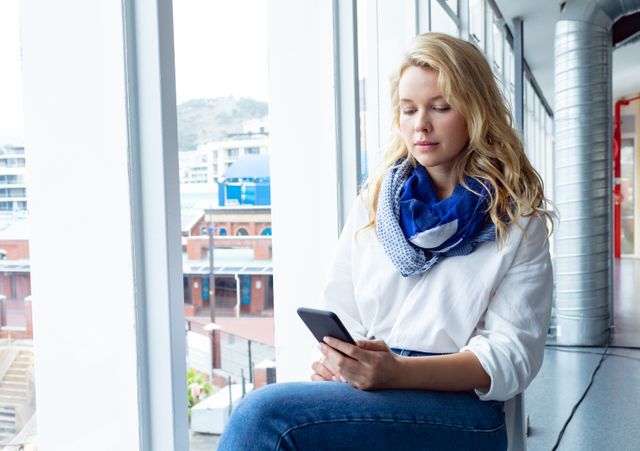 Front view of beautiful young Caucasian businesswoman using mobile phone near window in a modern office