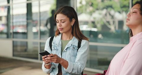 Diverse women waiting at city transit stop checking smartphone with backpacks