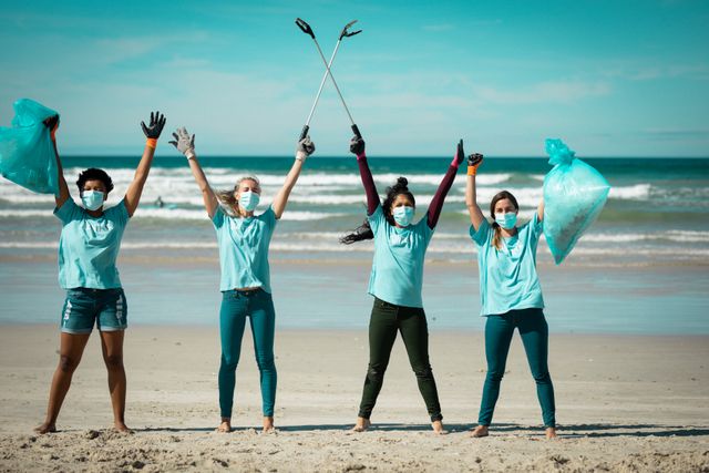 Diverse group of women wearing face masks and volunteer t-shirts picking up rubbish from a beach. They are holding trash bags and litter pickers, celebrating their efforts. Ideal for use in articles or campaigns about environmental conservation, community service, teamwork, and COVID-19 safety measures.