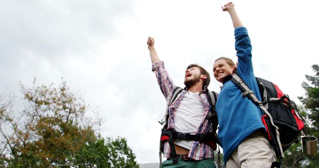 Two young Caucasian males are outdoors, one raising his fist in a triumphant gesture, both wearing backpacks and casual hiking attire. Their expressions and pose convey a sense of achievement and adventure, celebrating a successful hike or outdoor accomplishment.