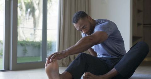 Man stretching hamstring on hardwood floor reaching foot near sliding glass doors in home