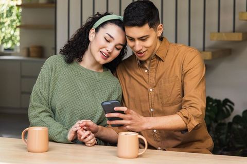 Asian couple sharing smartphone at cozy kitchen counter with coffee mugs and warm smiles