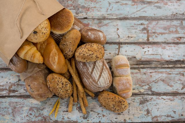 Assorted fresh bread loaves and rolls spilling out of a paper bag onto a rustic wooden table. Ideal for use in bakery promotions, food blogs, breakfast menus, and healthy eating campaigns. Highlights the variety and artisanal quality of the bread.