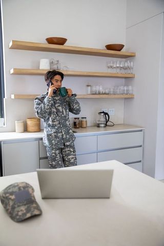 Asian service member in camouflage uniform talking on phone and drinking coffee in modern kitchen