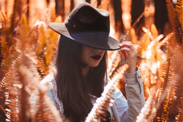Mysterious woman with long hair posing in autumn forest, wearing a black hat, surrounded by ferns. She holds the brim of her hat, adding to the enigmatic atmosphere. Ideal for articles on fashion, nature walks, autumn adventures, and mysterious character concepts.