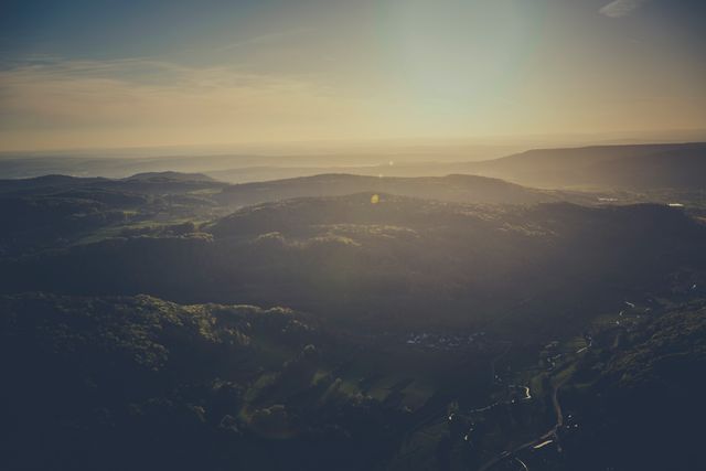 This image captures a serene aerial view of a hilly landscape at sunrise. The rolling hills are bathed in soft morning light, creating an atmosphere of peace and tranquility. The sun casts a gentle sunflare across the horizon, adding to the scenic beauty of the composition. Ideal for use in travel blogs, nature magazines, inspirational posters, or backgrounds for websites and digital content emphasizing natural beauty and calmness.