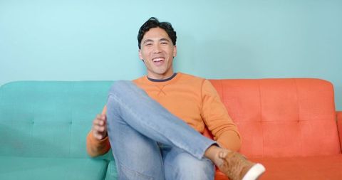 Smiling Man Sitting on Duotone Teal and Orange Sofa Wearing Orange Sweatshirt and Jeans