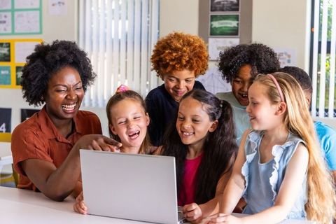 Sunlit Classroom Teacher and Diverse Children Laughing and Learning Together on Laptop