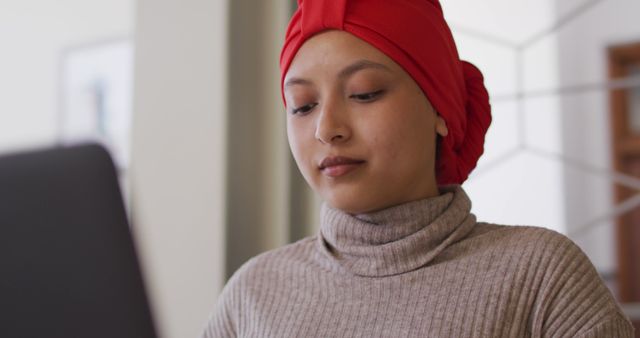 Young woman wearing red headscarf concentrated on work with a laptop in a home office. Perfect for themes related to remote work, productivity, professional settings, diverse work environments, and technology use by professionals.