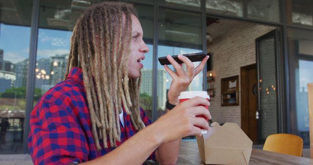 A man with long dreadlocks is using a voice command on his smartphone while holding a cup of coffee in an outdoor cafe. The relaxed atmosphere and modern setting make this image ideal for articles or advertising related to urban living, technology usage, convenience, or lifestyle blogs focusing on everyday activities.