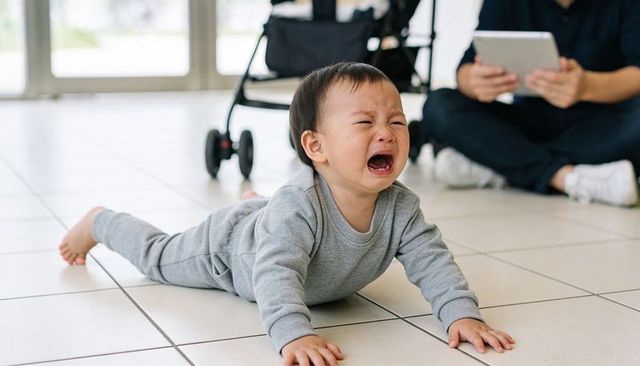 Crying toddler lying on tiled lobby floor near stroller while caregiver holding tablet