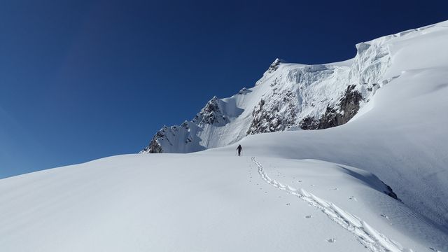 A solo hiker is trekking up a vast, snow-covered mountain with footprints trailing behind. Clear blue sky contrasts sharply with the pristine white snow. Ideal for travel, adventure, and outdoor activity themes. Suitable for articles, blogs, promotional materials on mountain climbing, winter sports, and exploration.