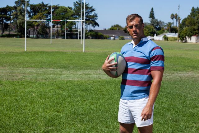 Portrait of confident rugby player holding ball while standing against goal post on field