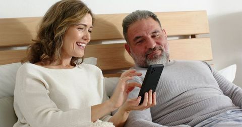 Reclining mature couple sharing smartphone in cozy bedroom, smiling and relaxing together