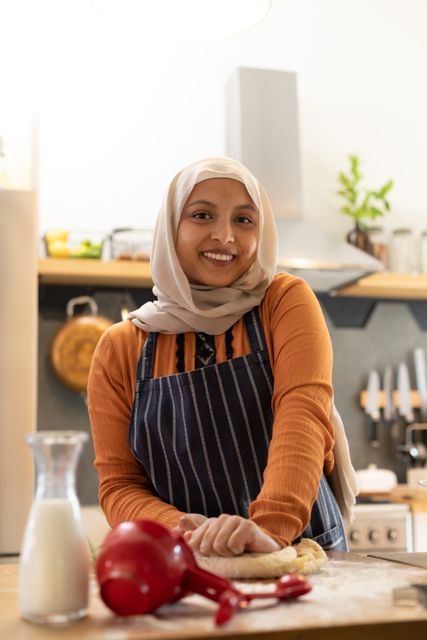Smiling woman in hijab kneading dough in a modern kitchen. Ideal for themes related to cooking, baking, domestic life, inclusivity, and multicultural representation. Perfect for use in articles, blogs, and advertisements promoting home cooking, family activities, and cultural diversity.