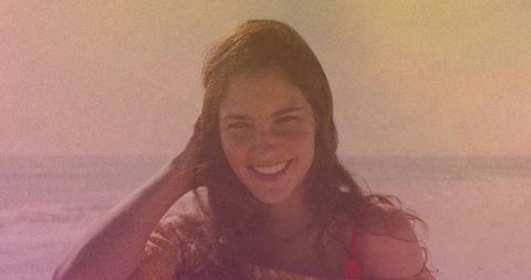 Smiling woman touching hair on sunlit beach wearing off-shoulder top with red strap