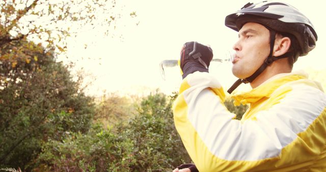 Male cyclist wearing a yellow jacket and helmet drinking water outside in a natural environment. Ideal for use in content promoting physical fitness, outdoor activities, sport hydration tips, cycling gear, or healthy lifestyle choices.