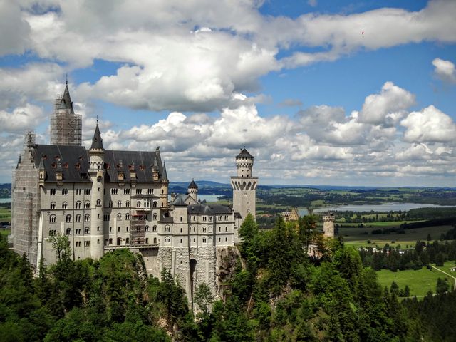 Neuschwanstein Castle stands majestically against a backdrop of lush green landscape and rolling hills under a partly cloudy sky. Ideal for travel brochures, historical articles, architectural studies, and touristic promotional material. Perfect for evoking a sense of fairytale wonder and historic grandeur.