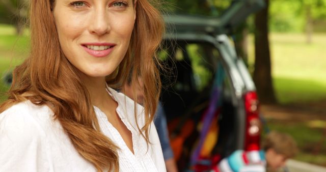 A woman is smiling near an open car ready for a road trip in a park with a green nature background. Ideal for travel, vacation, outdoor adventure, family activities, and advertisements promoting tourism.