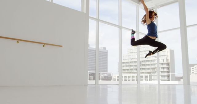 A young female dancer is seen mid-jump in a modern dance studio. The room has large windows which offer views of the urban skyline beyond. This image captures the skill, flexibility, and dynamic motion of a professional dancer. Great for advertising dance classes, promoting fitness programs, highlighting modern dance trends, and illustrating training and perseverance in sports and the arts.