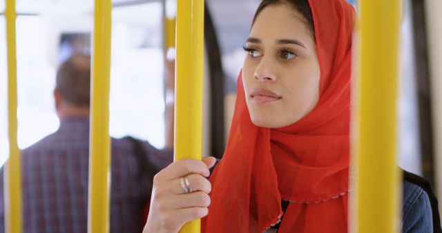 Young woman in a red hijab rides public transportation, with copy space. She appears contemplative while holding onto a yellow pole in a bus or train.