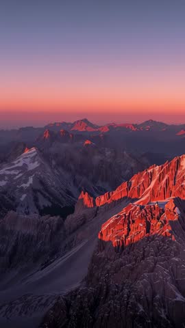 Vertical drone footage bathing alpine ridge in alpenglow at sunrise, casting red glow across peaks