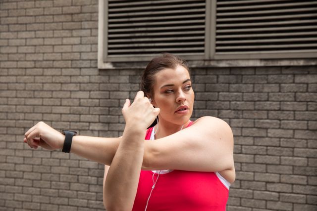 Curvy Caucasian woman with long dark hair wearing sports clothes and earphones exercising in a city, stretching her arms and warming up before her workout