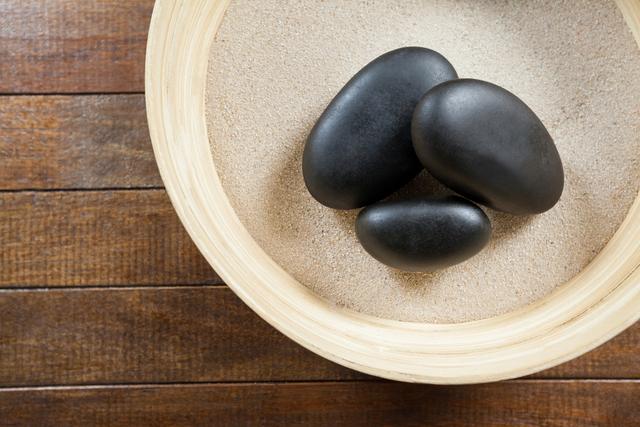 This image shows three smooth pebble stones placed in a wooden bowl filled with sand. The natural and minimalist composition evokes a sense of tranquility and balance, making it ideal for use in wellness, spa, and meditation contexts. It can also be used in designs related to relaxation, natural beauty, and simplicity.