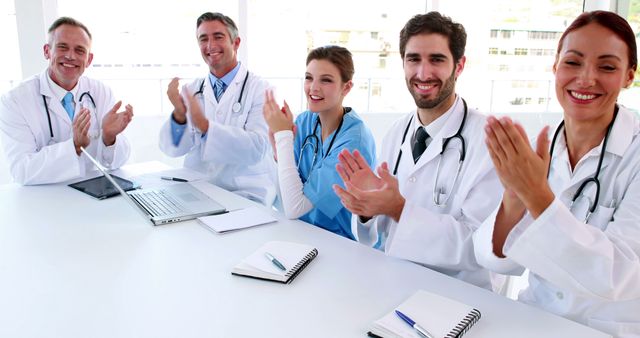 Several medical professionals, including doctors and a nurse, sit around a desk in a bright hospital room. They are applauding, smiling, and seem to be engaged in a discussion, suggesting a congratulatory or motivational atmosphere. Ideal for use in articles or advertisements about medical teamwork, healthcare success stories, collaborative healthcare efforts, and professional achievement in medical settings.