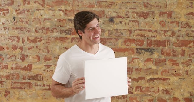 Portrait of happy caucasian man in white t-shirt holding white board with copy space by brick wall. Lifestyle, fashion and style, wellbeing, mock up, message and information, unaltered.