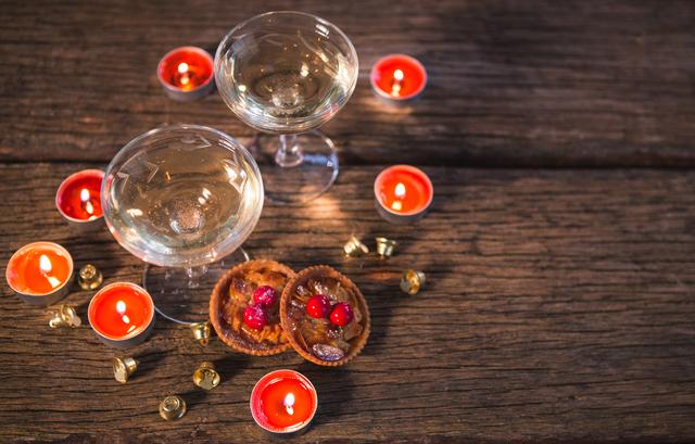 Two wine glasses are surrounded by red candles and festive decorations on a wooden table. Small desserts with berries add to the holiday atmosphere. This image is perfect for holiday greeting cards, festive blog posts, or social media content celebrating Christmas and the holiday season.