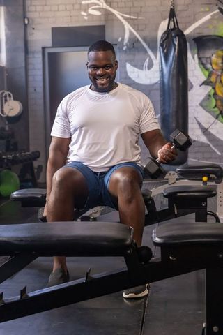 Smiling man doing seated dumbbell curl on bench in urban gym with graffiti wall