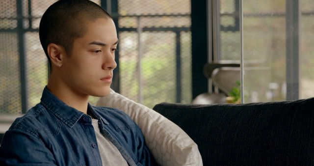 Young man is sitting on a couch indoors, looking thoughtful and pensive. He appears relaxed, dressed in casual clothing and sitting comfortably in a home environment. This image can be used for mental health campaigns, stock photos for articles related to contemplation and thought, or lifestyle blogs focusing on modern young adults.