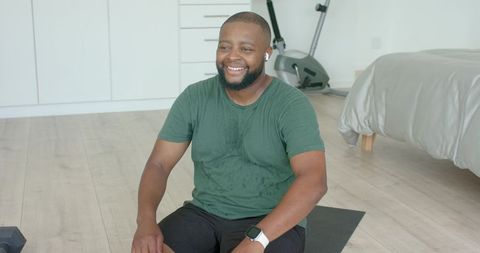 Smiling man sitting on exercise mat after home workout, sweating in minimalist bedroom gym