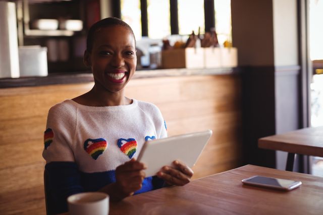 Woman sitting in a cafe holding a digital tablet and smiling. She is dressed casually in a white sweater with multi-colored heart patterns. There is a cup and a smartphone on the wooden table. Ideal for themes involving modern lifestyles, technology use in casual settings, and positive emotions.