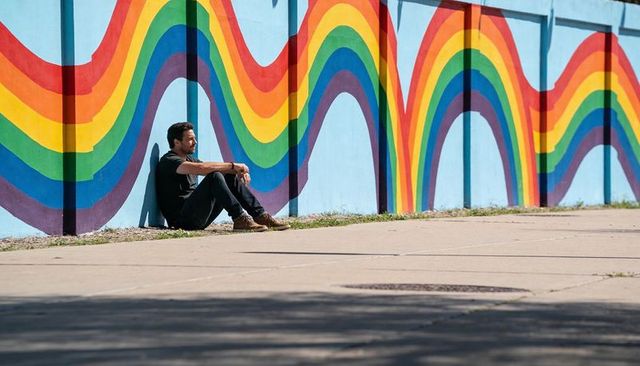 Man sitting against rainbow mural on sunlit urban sidewalk, contemplative solitary moment